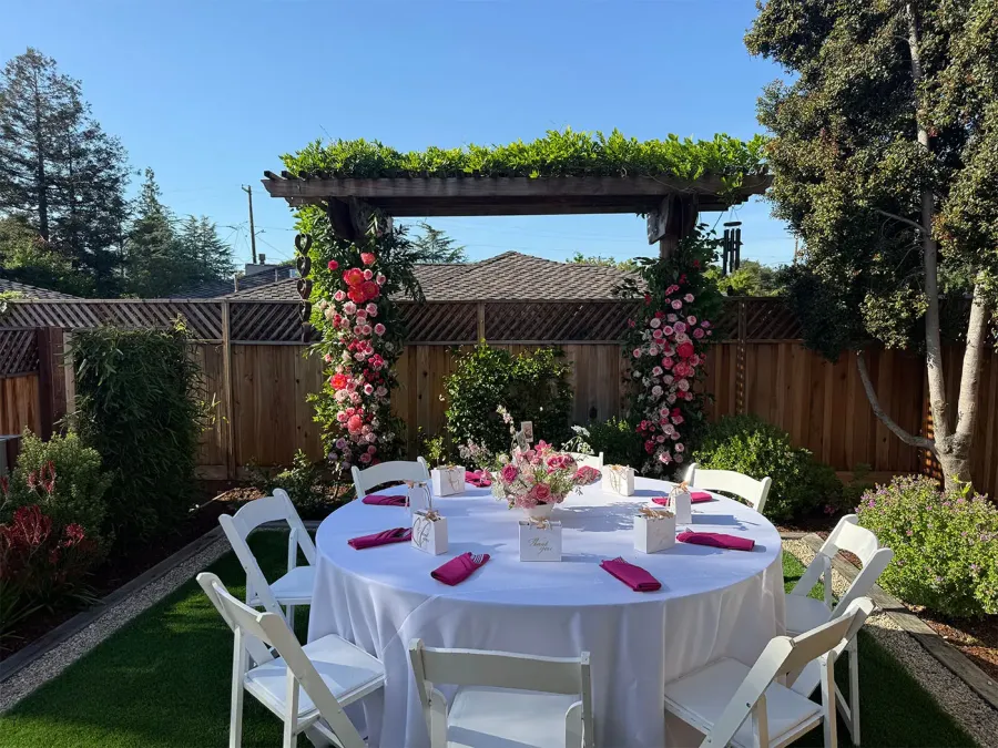 Rose arch in blush and deep red installed over a round garden table with white linen and fuchsia napkins — floral arch design for Marin catering weddings